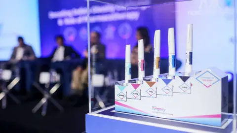 Bloomberg via Getty Images Five pens of weight-loss drug Wegovy jabs sit in an encased stand on a plinth in front of unidentifiable people sitting on chairs on a stage in front of a purple screen projection during a press conference for the launch of Wegovy in India, in Mumbai in June 2025.