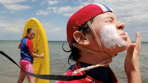 Getty Images A young child in a swimming cap applying sunscreen on their face, with another young child holding a surfboard, with the ocean in the background.