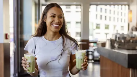 Getty Images Young woman buying matcha latte at coffee shop. Female holding two cold matcha latte glasses and walking inside city cafe.