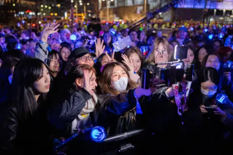 BIGHIT MUSIC AND NETFLIX A dozen fans looking at a camera with a light above it, surrounded by purple light-sticks