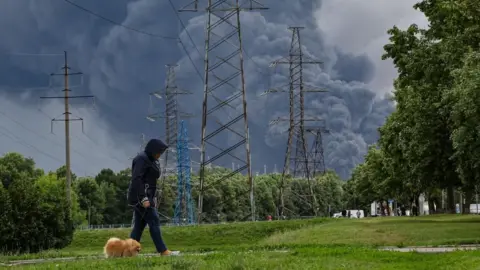 EPA A woman walks in a park as smoke billows in the background following a reported Ukrainian drone attack, in Moscow, Russia, 18 June 2026.