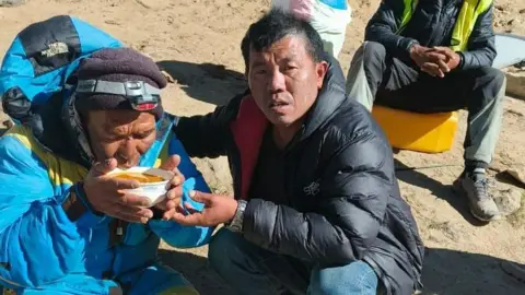 Sagarmatha Pollution Control Committee (SPCC) A man in a blue climbing coat drinking an orange soup from a bowl, being helped by a man in a black puffer coat