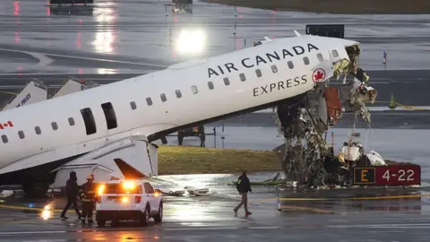 AFP via Getty Images Air Canada flight AC8646 with its nose upturned and wrecked. Emergency services surround it. The tarmac is wet, it is early morning. 