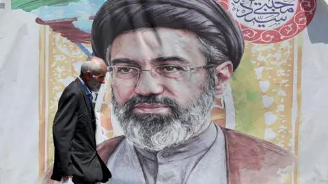 EPA A man with grey hair wearing a dark suit and blue shirt carrying a plastic bag walks past a poster of Supreme Leader Mojtaba Khamenei who has a grey beard and is wearing a grey buttoned shirt, maroon shawl glasses and a black turban.