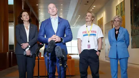 Getty Two women wearing pant suits and one man in a t-shirt look on as a man in a suit speaks into micoophones