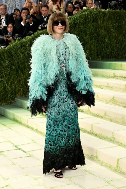 Getty Images Anna Wintour wears her hair in her signature bob and sports dark sunglasses. Her dress is an avian-inspired confection, with a turquoise feathered cape on top of a darker-green dress whose embroidery resembles feathers.