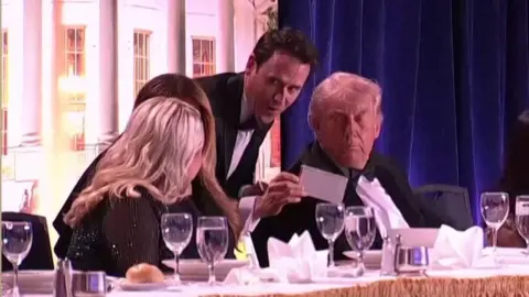 US pool Speaker Oz Pearlman (standing in the centre with a piece of paper) talks to US President Donald Trump (right), First Lady Melania Trump (2nd left) and White House press secretary Karoline Leavitt (left) at the White House Correspondents Dinner in Washington DC. Photo: 25 April 2026