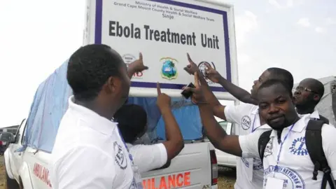 Patrick Faley Patrick Faley (L) and other community workers in Sinje point to a sign saying 'Ebola Treatment Unit' during the outbreak in Liberia