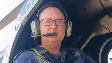 Johan Potgieter Captain Johan Potgieter sits in a helicopter, wearing his police uniform. He wears a pair of headphones with a microphone attached.