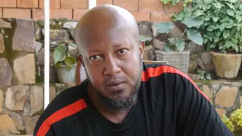 Ukuri Mbona via YouTube Close-up of Aimable Karasira, who is wearing a black T-shirt with red strips on the sleeves. He is sitting outside. There are potted plants and stone walls behind him