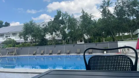 Emery Makumeno / BBC A large swimming-pool surrounded by a concrete wall with trees in the background