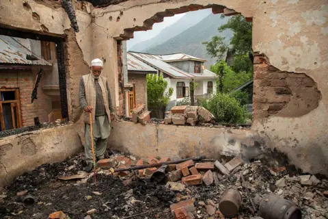 LightRocket via Getty Images An elderly man stands inside the burned-out shell of his home, leaning on a wooden walking stick and staring quietly at the debris around him. He wears a white skullcap, a grey sweater beneath a beige vest and loose traditional trousers. The room is gutted and blackened, its walls cracked.