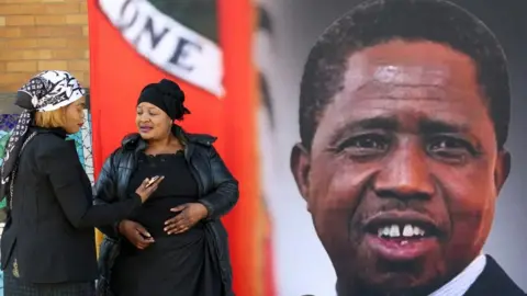 Reuters Mourners gather in front of a banner ahead of the funeral of Zambia's former President Edgar Lungu - the banner features a portrait of Lungu.