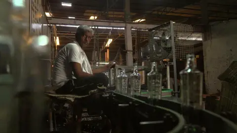Pritam Roy/BBC A worker - wearing a white undervest and a cotton scarf around his neck - sits next to a conveyer belt carrying glass bottles in a factory