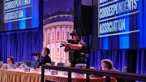 Getty Images an officer points a large gun as people duck behind a table beside him, with screens showing 
