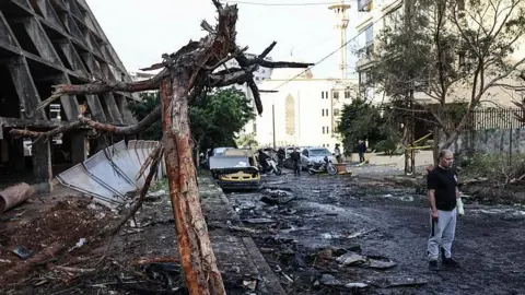 Getty Images A man stands in the street next to a building and tree which are both heavily damaged and blackened. Rubble lines the street and cars in the background are also damaged.
