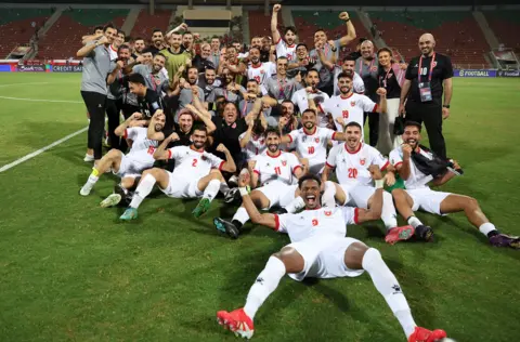 Ameen Ahmed/NurPhoto via Getty Images A group picture of Jordanian football players and other supporting members of the wider team smiling and celebrating on a football pitch. Many of the players are wearing white kit and sitting on the ground. They are all looking at the camera.