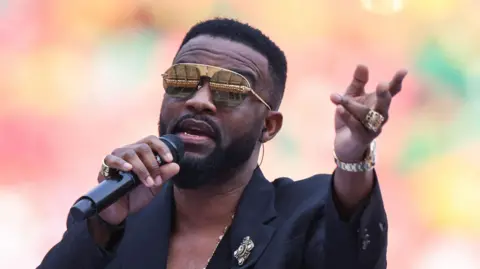 AFP via Getty Images Papa Wemba sings on stage wearing a black suit and large encrusted jewellery. In one hand he holds a microphone and with the other he reaches out.
