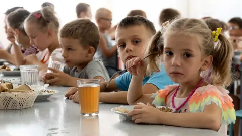 AP Children from an orphanage in the Donetsk region, eat a meal at a camp in Zolotaya Kosa, the settlement on the Sea of Azov, Rostov region, southwestern Russia, Friday, July 8, 2022. A little girl with plaits is on the right, closest to the camera, she is looking sadly at the camera and holding a fork in her right hand over her plate of food. A glass of red-brown liquid is on the table beside her. A boy next to her is also looking at the camera. Other children are busy eating and drinking.