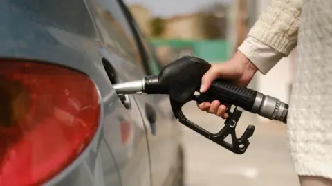 Getty Images A woman's arm and hand holding a black petrol pump and using it to fill her silver car