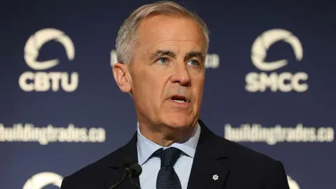 Getty Images An image showing Carney speaking at a trades event. He has short grey and black hair and blue eyes. He is wearing a black suit with a light blue shirt and a navy blue dotted tie.