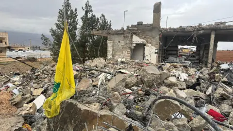 BBC The yellow flag of the Hezbollah group hanging by the ruins of a house