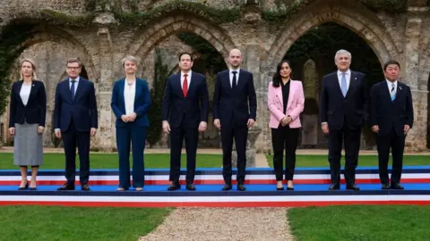 Reuters A row of the leaders stood in a line facing forward and smiling. They're on a short stage and are dressed smartly. A fancy old stone building with archways is in the background