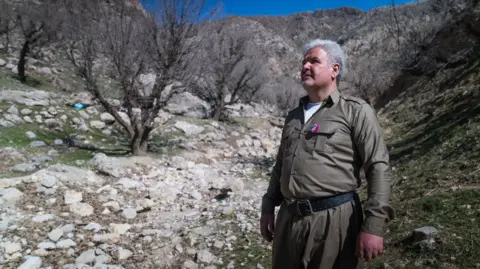 Matthew Goddard/BBC Shaho Bloori, dressed in grey uniform, stands to the right of the picture looking up and to the left in a barren mountainous landscape with a few small trees