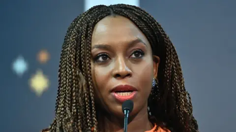 Getty Images Chimamanda Ngozi Adichie talks into a microphone. She wears an orange top and long braids.