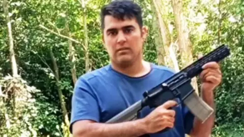 Noah Aaron stands outdoors in a wooded area with dense green trees and sunlight filtering through the foliage. He is wearing a short-sleeved blue T-shirt and is holding a long firearm with both hands; the firearm has a visible magazine and is angled slightly across the body. The background consists of tree trunks, leaves, and undergrowth, with no buildings or other people visible.
