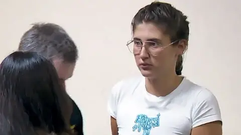 Rustavi 2 Bella Culley, in a white-walled court room, although she is seen close up so her surroundings are not shown. She is wearing silver-rimmed aviator glasses and has brown hair in a french plait. Her t-shirt is short sleeved and white with a turquoise design on the chest. She looks very apprehensive. In front of her are two people - one seen from behind and the other obscured by the first.