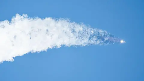 Getty Images A space rocket flies upwards into a clear blue sky, leaving a huge grey trail behind it in a widening streak.
