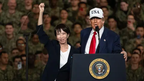 Getty Images Sanae Takaichi (left) punches fist into the air as Donald Trump, wearing a white cap that says USA, speaks in front of US Navy personnel on board the USS George Washington aircraft carrier in Japan