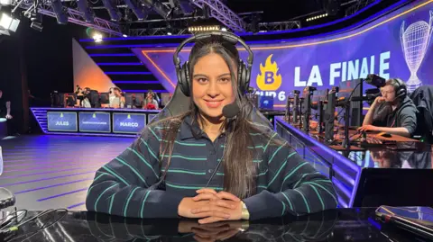 BBC A young woman is sitting in a computer chair, in an e-sport venue. She is looking directly into the camera, smiling. Her hair is dark and long, she is wearing a striped shirt and a headset. Behind her is a large screen displaying the word 'final' in French, and a row of chess players in front of computer screens. 