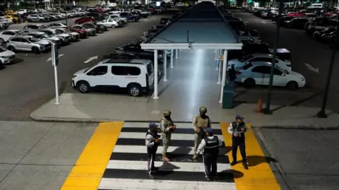 REUTERS/Cesar Munoz Members of Ecuador's armed forces stand stand guard after an armed attack at the airport in Guayaquil. There are two soldiers with guns and three other uniformed men standing on a pedestrian crossing leading to the parking lot. 