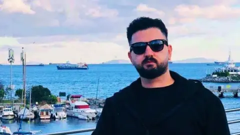 BBC Kardo Jaf pictured in a coastal setting. He has short black hair and facial hair, and wears black sunglasses and a black top. Behind him is a large body of water where several boats are docked in a marina, and 