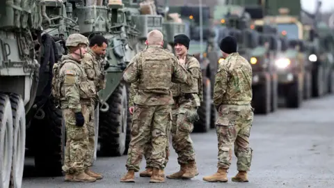 Getty Images Six US soldiers in green camouflage uniforms are pictured in Germany standing in a circle, with a long line of military vehicles visible behind them.