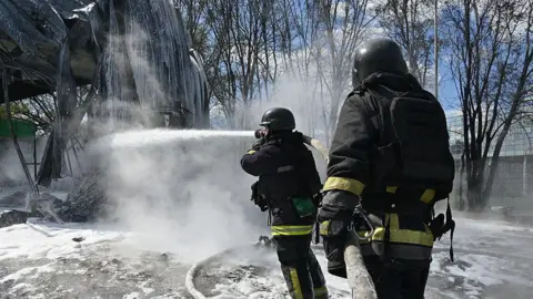 Getty Images Two Ukrainian firefighters hold a hose as they try to extinguish a fire at a destroyed petrol station in Kharkiv - 2 May.