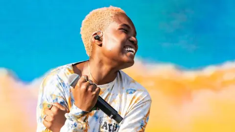 Getty Images Arlo Parks gives a disarmingly carefree smile as she performs in Australia. She's holding a microphone in her right hand, and wearing a colourful long-sleeved t-shirt, whose blue and yellow colours are reflected on the stage behind her.