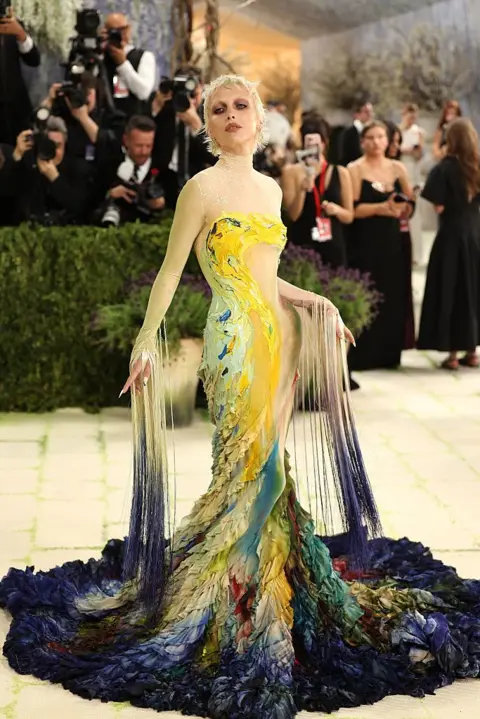 Getty Images Influencer Emma Chamberlian wears a multi-colored gown that resembles a painting. It begins with dark blue colors at the base before transforming into a yellow and teal abstraction. Her sleeves also have fringes down to the floor.