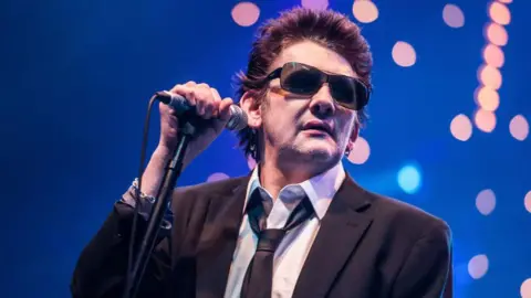 Getty Images Shane MacGowan is pictured in tinted sunglasses and holding a microphone, while preparing to sing at a concert in 2012. The background is a dark blue stage backdrop with a bloom of spotlights.