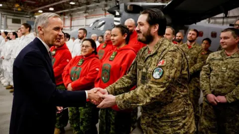 Reuters Prime Minister Mark Carney is seen shaking hands with a service member of the Canadian Armed Forces. Behind them are rows of service members from different units, standing in front of a military aircraft inside a hangar.