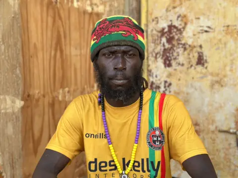 Brandon Drenon / BBC Roy Perry wearing a yellow t-shirt seated in front of a badly damaged building