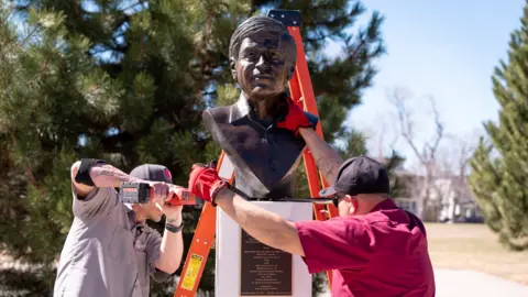 Reuters Two men with power tools remove a bust of Cesar Chavez from a pedestal. The men wear black baseball hats. 