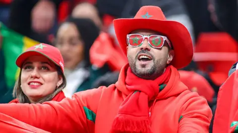 Getty Images Two Moroccan fans - a man and a woman - side-by-side in the stadium. The man is wearing a cowboy hat in Moroccan colours and sunglasses with the Moroccan flag on them. The woman is wearing a Morocco baseball cap.