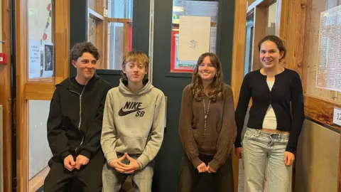 Four teenagers stand smiling in a school corridor - two boys and two girls. They are dressed casually