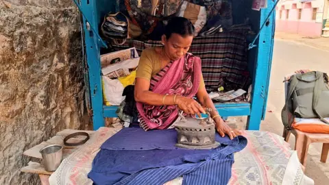 NurPhoto via Getty Images A woman wearing a pink sari irons a blue shirt at her roadside shop in Odisha, India.