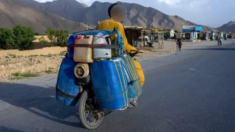 BBC Mazaar is riding his small motorbike with canisters filled with fuel on the back of it. He's on a wide road in a semi-rural area with mountains in front of him and a few vehicles