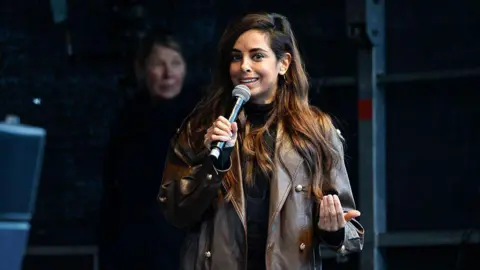 Morris MacMatzen/Getty Images A woman in a brown leather jacket and brown hair holds a microphone 