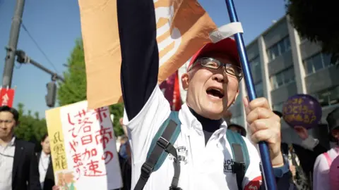 BBC News: Jiro Akiba A man in a backpack, glasses and a red cap punches a hand in the air while holding a flag